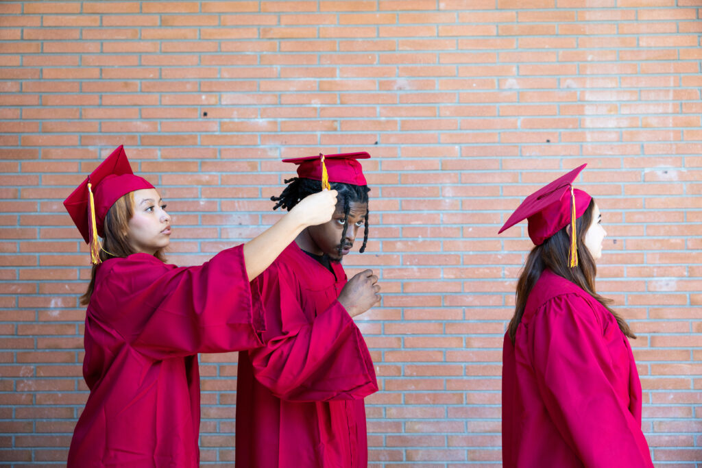 Two female students in graduation cap and gown adjusting the mortarboard of a male student in preparation for a graduation ceremony on a college campus.