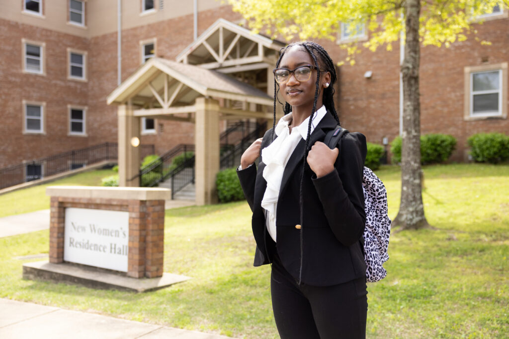 Black female student in a suit and glasses posing for a portrait in front of a residence hall on a college campus.